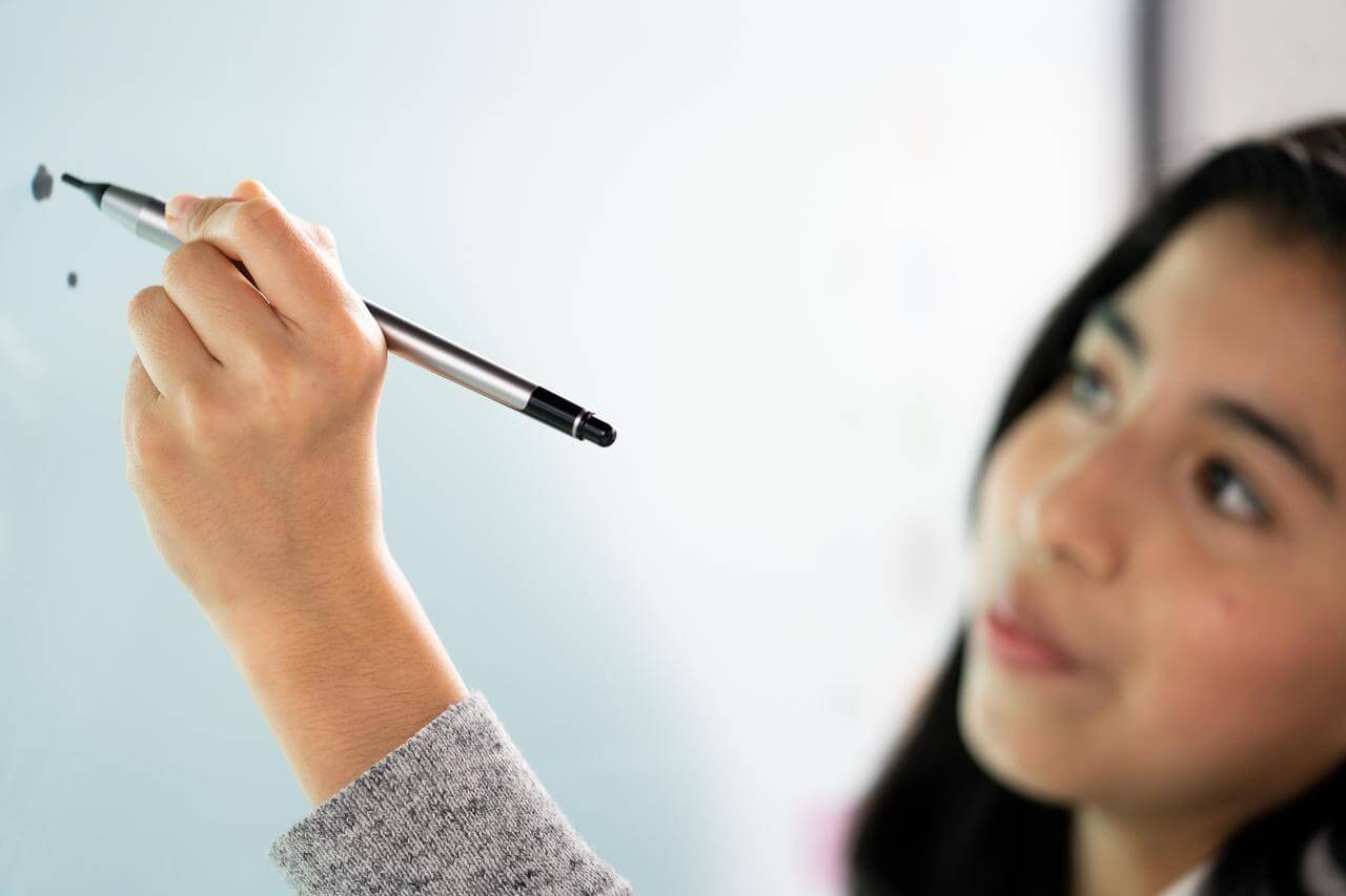 A school child stands in front of an interactive whiteboard, holding a stylus in their hand, concentrating on the start of a drawing.